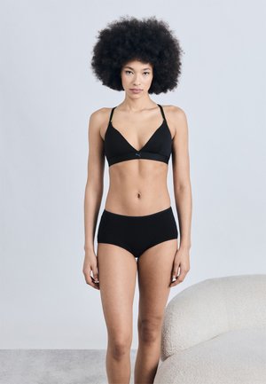 Γυναίκα με αφροχτένισμα, που φοράει μαύρο αθλητικό σουτιέν και μαύρο σορτς, στέκεται δίπλα σε μπεζ καμπυλωτό καναπέ μπροστά σε απλή ανοιχτόχρωμη τοιχοποιία.