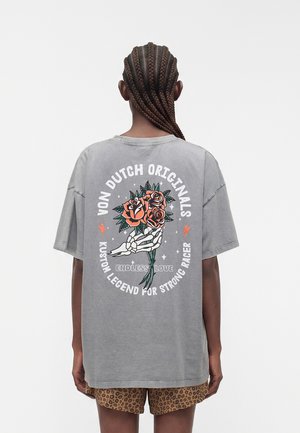 Person mit geflochtenem Haar trägt ein graues oversized T-Shirt mit einem floralen Skelett-Hand-Grafik und Text, kombiniert mit Shorts mit Leopardenmuster.
