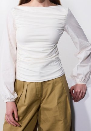 Vrouw draagt een witte blouse met lange, gerimpelde mouwen en pofmouwen, en een beige broek met hoge taille en wijde pijpen, staand tegen een witte achtergrond.