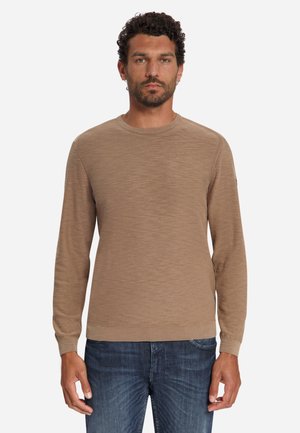 Man with curly hair and beard wearing a light brown long-sleeve sweater and blue jeans standing against a plain background.