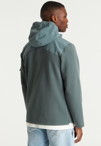 Gewatteerde jas in een tweekleurig ontwerp; donkergroen lichaam met een lichtere teal kap en schouderpaneel. Zachte textuur met minimale hardware.