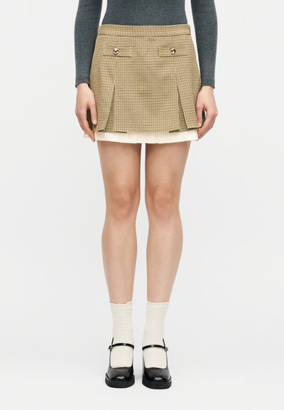 Femme portant une mini-jupe plissée à carreaux beige superposée sur une jupe blanche, associée à des chaussettes blanches et des chaussures Mary Jane noires.