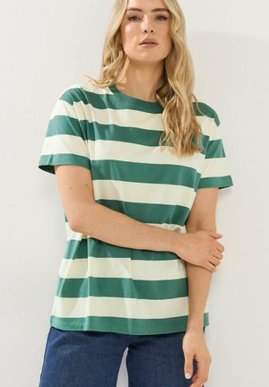Blonde Frau in einem kurzärmligen T-Shirt mit grünen und weißen horizontalen Streifen und blauen Jeans, die mit verschränkten Armen steht.