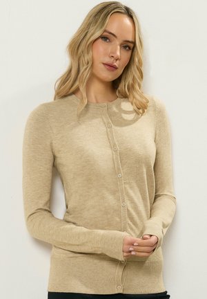 Blonde Frau, die eine beigen, geknöpften Strickjacke mit langen Ärmeln trägt, vor einem schlichten hellen Hintergrund steht.