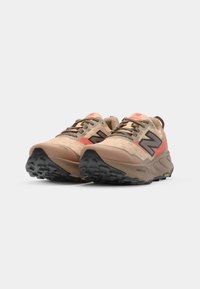 Des chaussures de trail de couleur beige avec une tige en mesh, un logo noir, des accents rouges et des semelles extérieures en caoutchouc texturé pour l'adhérence. Col rembourré pour le confort.