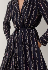 Napapaitettu mekko tummansinisestä kankaasta, jossa on kermavalkoinen ja pastellivärinen ketjukuosi. Pitkät hihat ja vyötäröllä solmittava yksityiskohta.