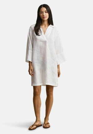 Femme aux longs cheveux noirs portant une robe blanche ample mi-longue avec des manches larges et des sandales plates marron, debout sur un fond uni.