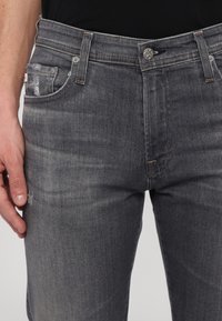 Homme portant un jean noir délavé avec une légère usure sur la poche avant et une chemise noire rentrée.