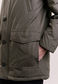 Kurtka ocieplana w oliwkowej zieleni, z dużymi przednimi kieszeniami, zapięciem na guziki i przeszyciami po bokach. Gładka faktura materiału.