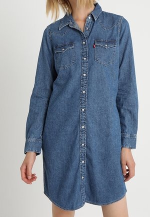 Kvinde iført en langærmet blå denimskjortekjole med knapper foran og to brystlommer mod en ensfarvet baggrund.