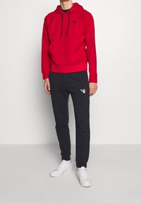 Red cotton hoodie with a black logo, paired with navy sweatpants featuring a multicolored logo. White sneakers complete the outfit.