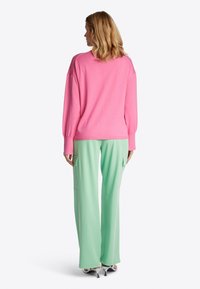 Rosa Strickpullover mit überschnittenen Schultern, kombiniert mit mintgrünen, weiten Hosen mit seitlichen Taschen. Das Model wird von hinten betrachtet.