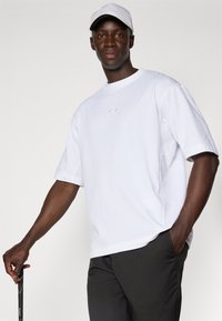 Hvid bomulds t-shirt med korte ærmer, prydet med et diskret logo i midten. Kombineret med mørke bukser og en hvid kasket.