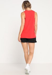 Czerwona bezrękawnik z okrągłym dekoltem, gładką teksturą i prostym wykończeniem, w połączeniu z czarnymi szortami i białymi butami sportowymi.