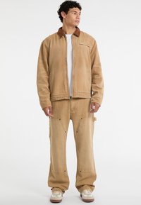 Beige Canvas-Jacke mit einem braunen Cordkragen, kombiniert mit passenden Cargo-Hosen mit großen Taschen und Metallakzenten.