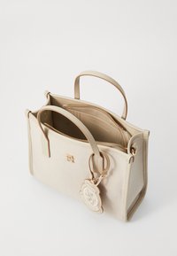 Bolsa bege feita de material suave, com duas alças, um fecho dourado e um charme decorativo com um brasão. O interior possui dois compartimentos.