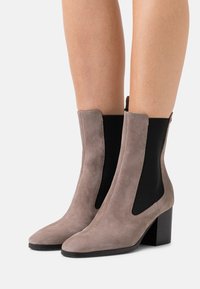 Bottes en suede taupe hauteur cheville avec des panneaux latéraux élastiques noirs et des talons blocs portées sur des jambes nues sur un fond blanc uni.