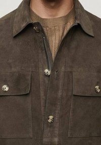 Uomo che indossa una giacca in suede marrone sopra un maglione beige a coste, con tasche a pattina con bottone e una zip frontale parzialmente aperta.