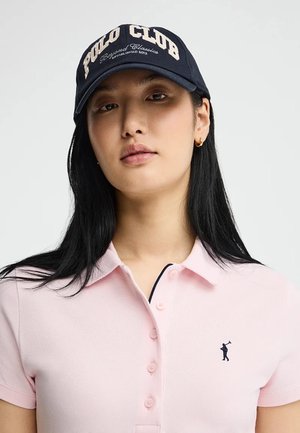 Joven mujer con una gorra azul marino que dice "Polo Club" y una camisa polo rosa claro con un pequeño logo de golfista en el pecho.