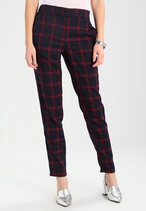 Frau trägt marineblaue Hose mit rotem Karomuster und silbernen Blockabsatzschuhen, steht mit locker neben dem Körper hängenden Händen.