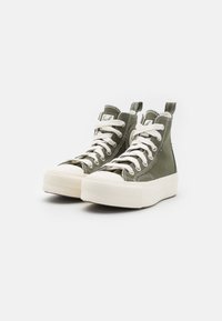 Vysoké tenisky z olivově zelené plátěné tkaniny s bílými tkaničkami a gumovými podrážkami. Mezi vlastnosti patří texturovaná, vzorovaná špička a poutko na patě.