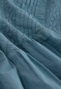 Blauwe stof met een zachte textuur, voorzien van verticale strepen en ingewikkelde kantdetails. Geplooide secties voegen diepte en dimensie toe.