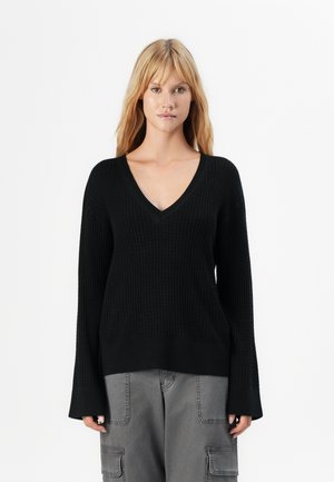 Woman with long blonde hair wearing a black V-neck knit sweater and gray cargo pants, standing against a white background.
