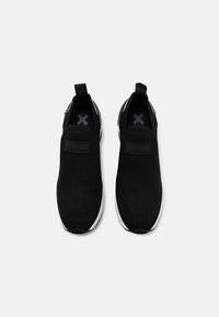 Scarpe slip-on in rete nera con una superficie testurizzata, dotate di un elastico con logo e suola bianca imbottita. Due linguette posteriori per indossarle facilmente.