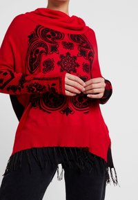 Rode trui met een col en zwart geborduurde patronen. Voorzien van een franjeszoom en geribbelde boorden. Gemaakt van zacht gebreid materiaal.