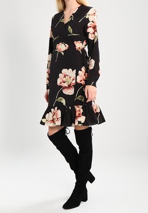 Kobieta ubrana w czarną sukienkę w kwiaty z długim rękawem i falbaną na dole oraz czarne zamszowe kozaki za kolano, stojąca na tle jednolitego tła.