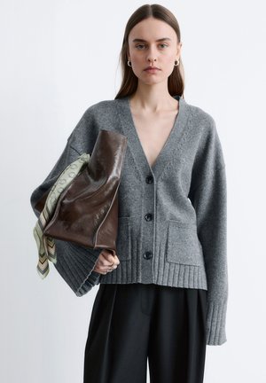 Kobieta w szarym, zapinanym na guziki kardiganie i czarnych spodniach, trzymająca dużą brązową skórzaną torbę, z której wisi wzorzysty szal.