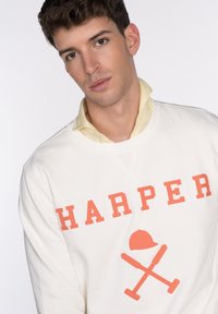 Sudadera blanca con la inscripción "HARPER" en letras color coral y un gráfico de herramientas cruzadas y un casco. Llevada sobre una camiseta amarilla clara.