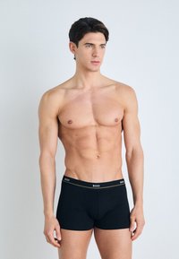 Caleçons boxers noirs avec une ceinture arborant un logo. Le tissu semble lisse et élastique, conçu pour le confort et l'ajustement.