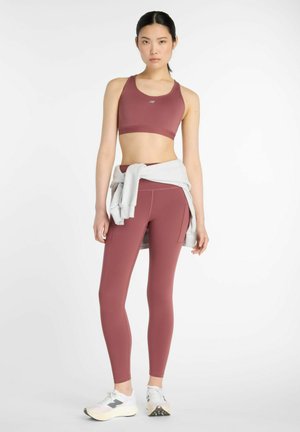 Giovane donna in piedi che indossa un reggiseno sportivo mauve e leggings, scarpe da ginnastica bianche, con una felpa grigia legata attorno alla vita su uno sfondo bianco puro.