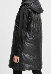 Czarna pikowana kamizelka z kapturem, o pikowanej strukturze, bocznych zamkach błyskawicznych i połyskującym wykończeniu. Krój jest długi i luźny.