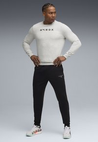 Sweatshirt gris avec le logo "HYROX", associé à un pantalon de jogging noir arborant le même logo. Modèle portant des baskets blanches avec des accents multicolores.