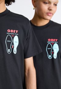 Tricouri negre din bumbac cu un text grafic îndrăzneț "OBEY alternative facts" și urme de pantofi stilizate, una albă și una neagră, etichetate R și L.
