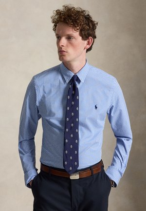 Homme aux cheveux bouclés portant une chemise à carreaux bleue, une cravate bleu marine avec des motifs de blason, une ceinture marron et un pantalon foncé, debout les mains dans les poches.