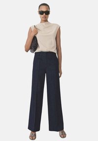 Beige ärmlös topp med hög hals, tillsammans med marinblå vida byxor med framveck; modell som håller en svart texturerad väska.