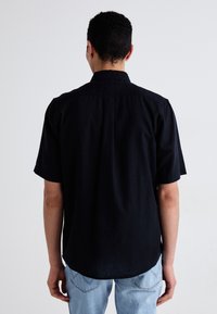 Maglietta nera a maniche corte con vestibilità rilassata, orlo dritto e colletto, realizzata in tessuto morbido e con una leggera lucentezza, indossata sopra jeans azzurri chiari.