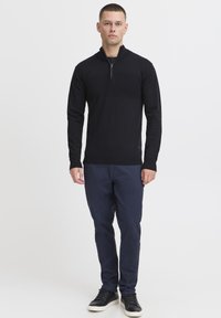 Svart glidelås-genser i en tettsittende design, laget av mykt materiale. Kombinert med marineblå bukser og svarte sneakers. Enkel og funksjonell.