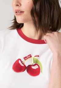 Biała koszulka z krótkim rękawem i czerwonym kołnierzykiem, z grafiką dwóch czerwonych wiśni z zielonymi liśćmi oraz małą czerwoną metką nad wiśniami.