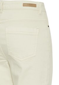 Pantalones de mezclilla color blanco roto que muestran el bolsillo trasero, las trabillas del cinturón y la etiqueta de la marca beige encima del bolsillo en la cinturilla.