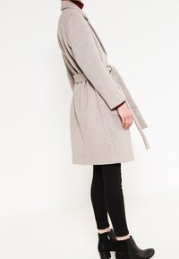 Beige wolmix jas met een riem, reverskraag en lange mouwen, gecombineerd met zwarte leggings en enkellaarsjes. Gladde textuur, knielang.