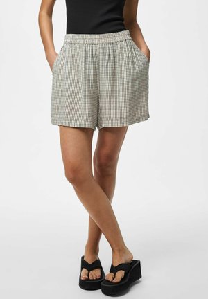 Nő bézs kockás rövidnadrágban és fekete ujjatlan felsőben, zsebre tett kézzel, keresztbe tett lábakkal, fekete platform szandálban áll.