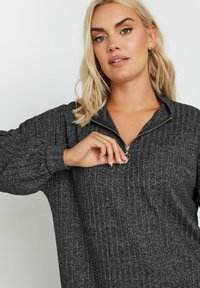 Maglione grigio con zip, texture a coste, cerniera argentata e spalle scese. Il tessuto appare morbido; presenta un colletto alto e una vestibilità rilassata.