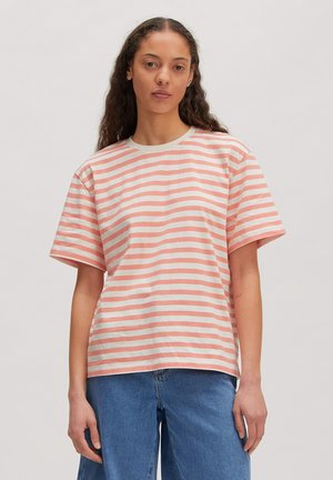 Frau mit langen lockigen Haaren, die ein rosa-weiß gestreiftes kurzärmeliges Shirt und blaue Jeans vor einem schlichten Hintergrund trägt.