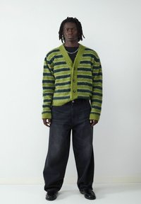 Sweter w kolorze zielonym i ciemnoniebieskim w paski, z dekoltem w serek i zapięciem na guziki, noszony na czarnej bluzce, w połączeniu z szerokimi ciemnymi dżinsami.