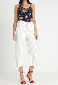 Canotta nera con motivo floreale e pizzo, culotte bianche a righe e tacchi a strappo rossi. L'outfit presenta una vestibilità ampia e una lunghezza alla caviglia.