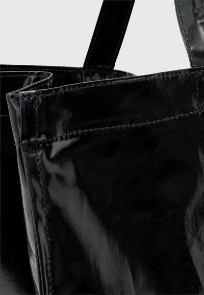 Gros plan d’un sac cabas noir brillant montrant des coutures détaillées et deux larges anses sur un fond uni.